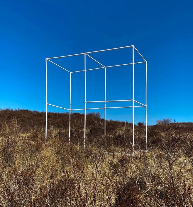 A thin white metal cuboid frame stands on one end in an overgrown field in front of a clear blue sky.