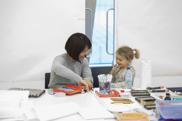 a dark haired woman sits next to a young girl in an art workshop; they are laughing and chatting and clearly are from different generations and ethnic heritages 
