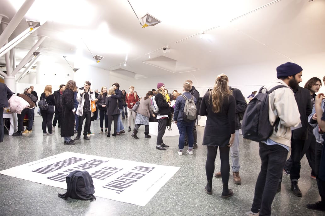 A large, crowded gallery space filled with people, with a flat, floor-based fabric work in the foreground
