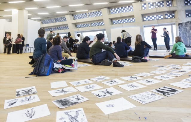 groups of people of varying ages, mostly seated on floor cushions, observe Rachel Gadsden as she leads n art workshop in front of a large blackboard with florid sketches on it