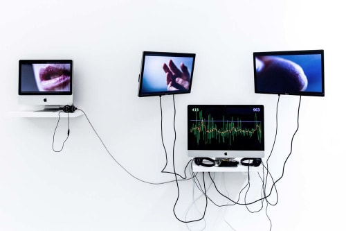 Image shows four computer screens against a white wall, two screens show close ups of mouth and hands and the others show the rate of the heart beat