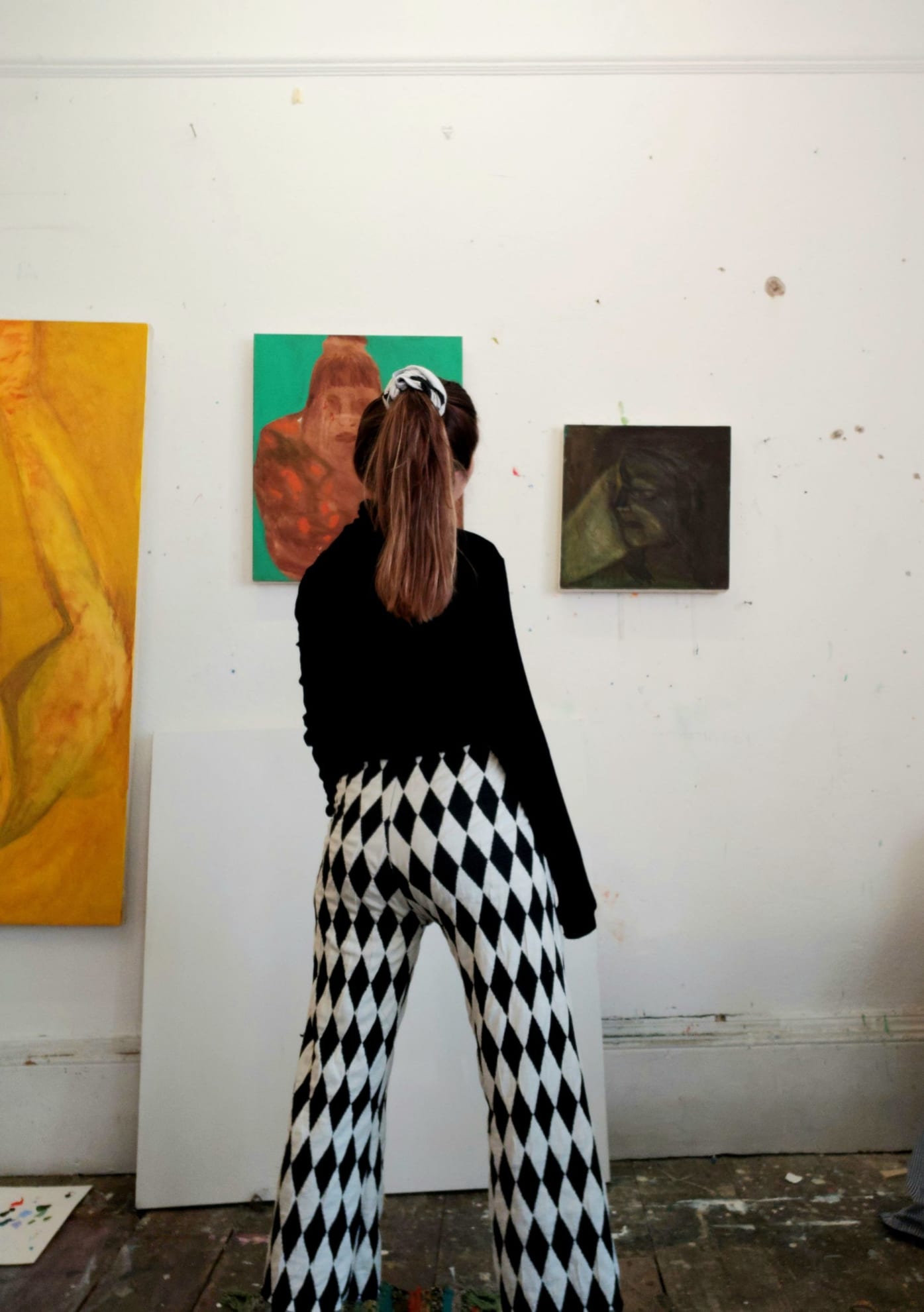 Oriele stands facing the wall of a studio. Her paintings line the wall in a row of three. Oriele is wearing black and white, boldly patterned trousers and black top. Her hair is tied up in a ponytail. 