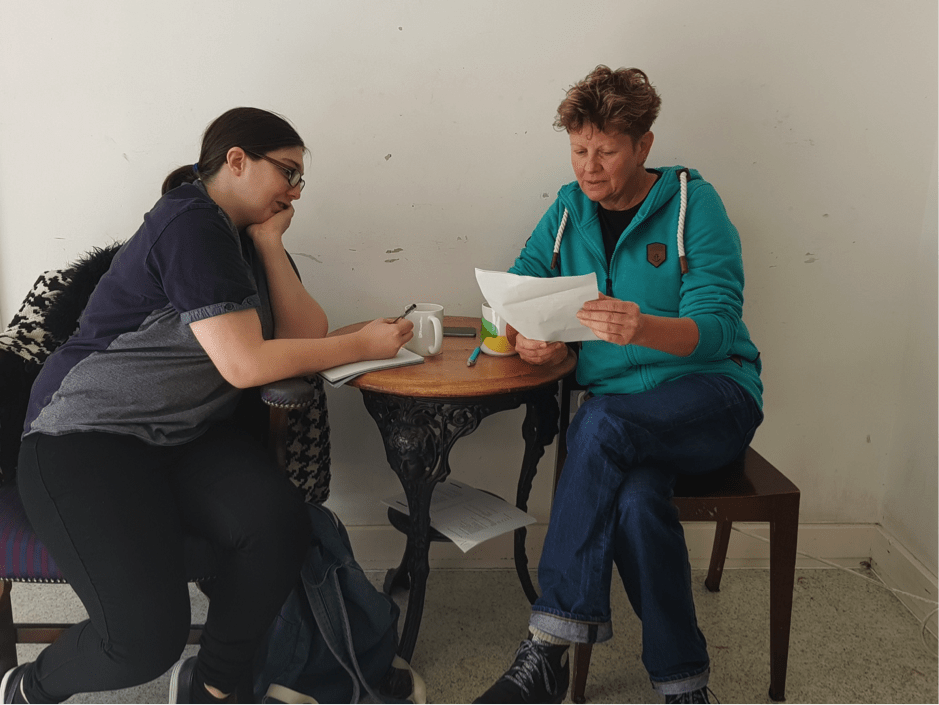 Two white women sit inside at a small table in front of a white wall. They are looking at a piece of white paper together with neutral expressions