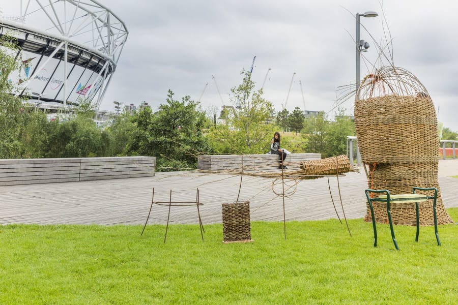 A collection of medium-sized sculptures woven from willow are standing in a park; there is a stadium in the background