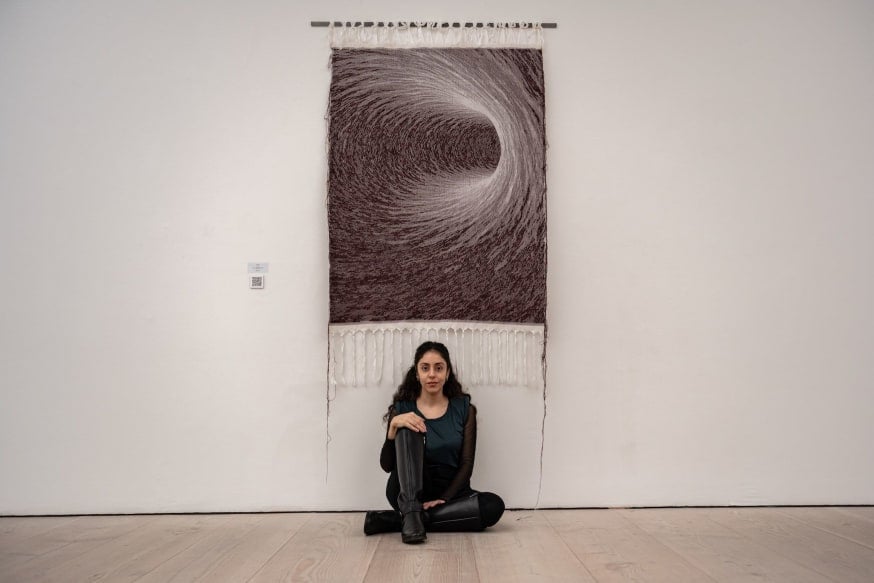 Hung on a long white wall is a brown and white swirling tapestry titled ΞXIT by MH Sarkis. Sarkis sits below the tapestry leant against the white wall. One knee is raised and Sarkis sits in a relaxed pose.