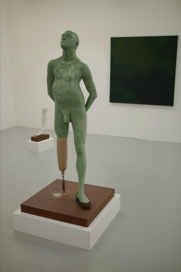 A green, realistic. life-size sculpture of a man with a wooden prosthetic leg is standing on a wooden plinth in a white-walled gallery room. The man is naked and his pose is defiant and proud, with his hands lightly clasped behind his back.