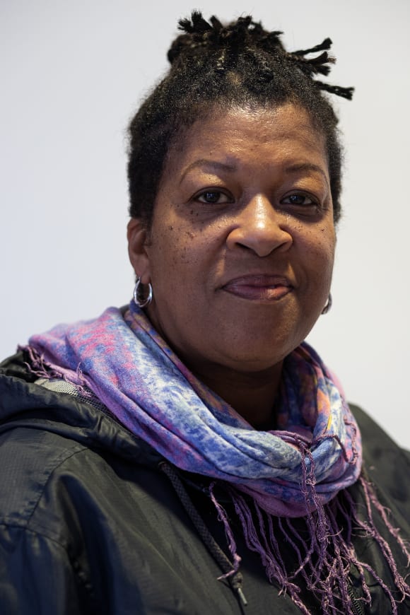 This headshot portrait of Suzie shows a black woman in her 60s facing the camera from the left with a relaxed, calm expression. Her hair is dark and tied up in braids on top of her head. Over a dark hooded coat or jacket she wears a purple and blue s