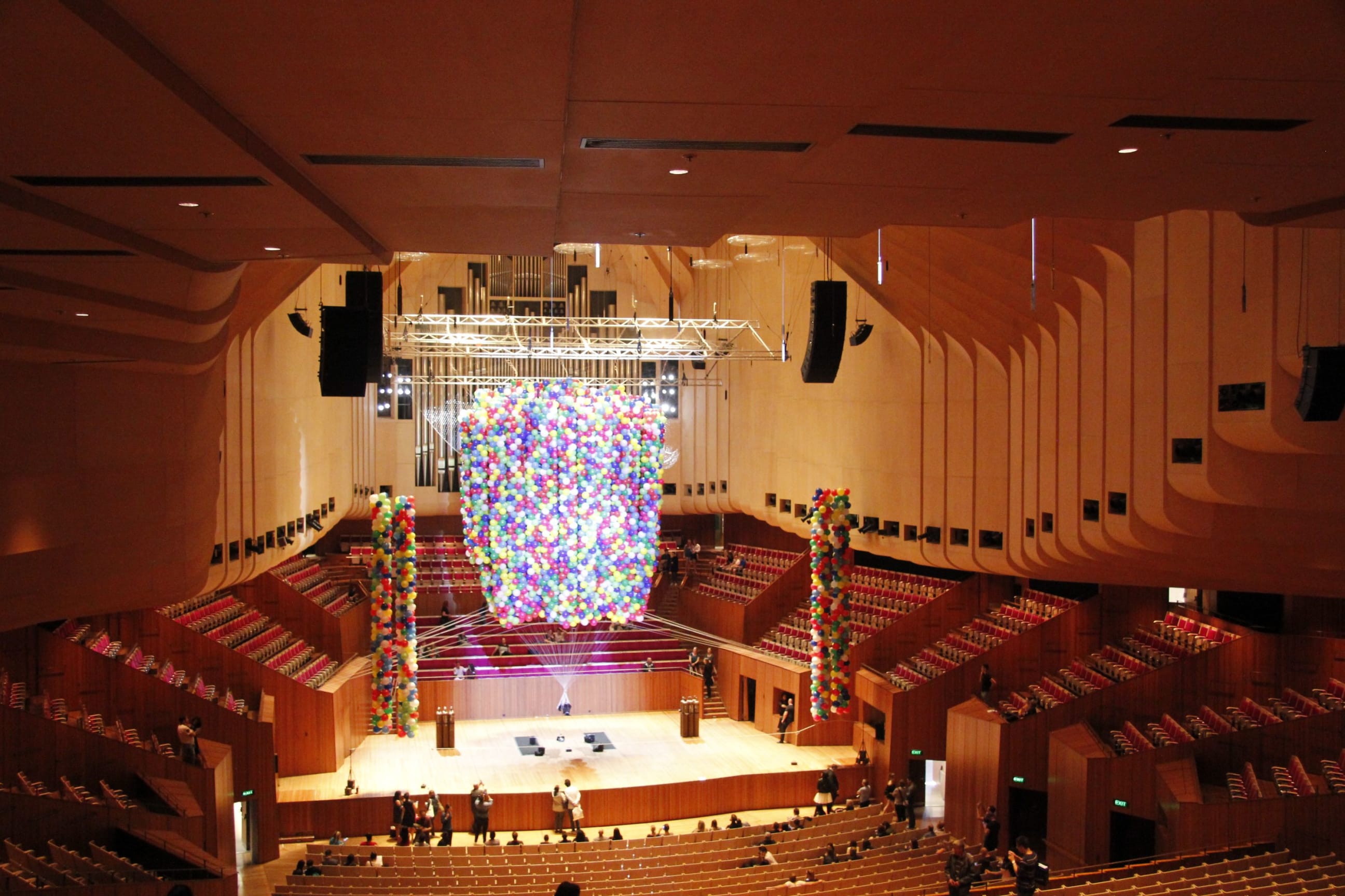 A vast, empty, wood-panelled concert hall (the Sydney Opera House) with a huge bunch of multicoloured helium balloons floating together above the stage with a very small human figure suspended laying down at the bottom of them