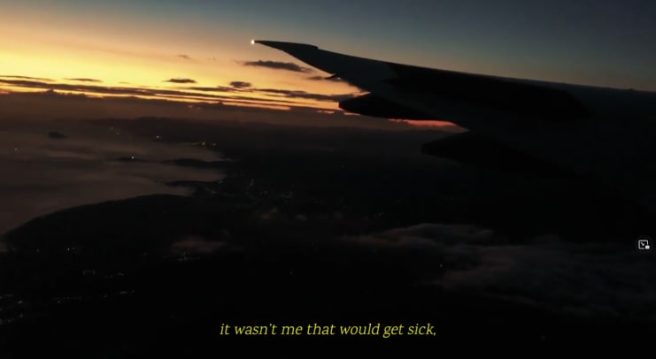A view out of a plane at sunset or sunrise. Light is low in the sky and there is an outline of the left wing of the plane. There are clouds below across the sky. Yellow subtitles read: 