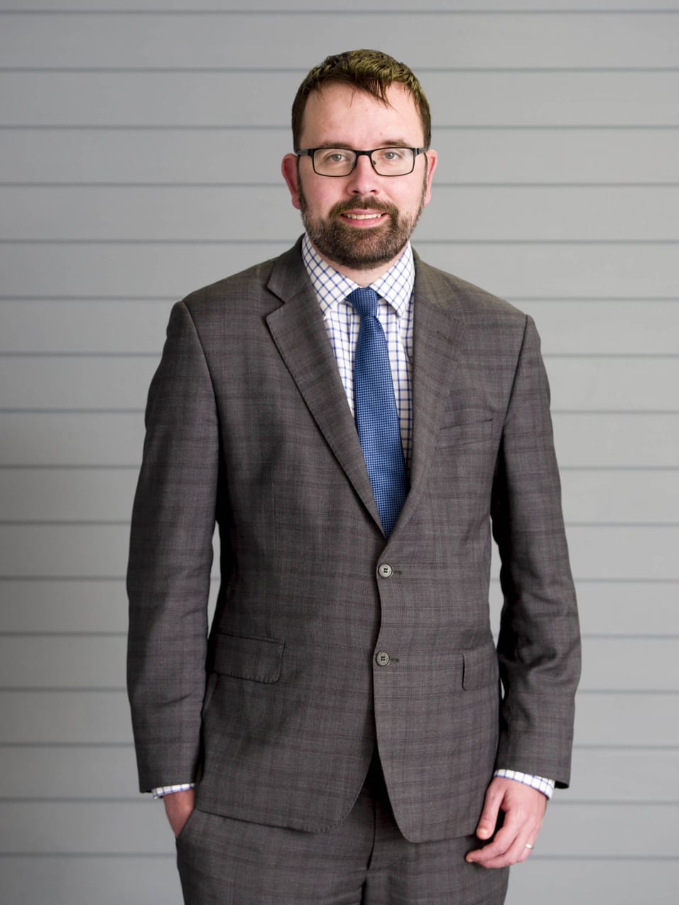 Photo of a man wearing a grey suit, white shirt, and blue tie. He has short brown hair and a short brown beard. He wears dark glasses and has one hand in a pocket.