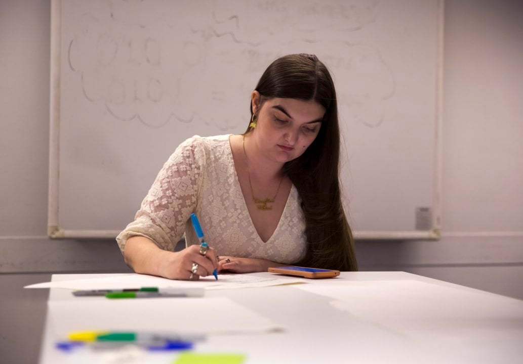 Photograph of artist Charlie Fitz, a young woman with long brown hair falling down past her shoulders. She is wearing a lacy white dress with a v-neckline and three-quarter length sleeves. She is sat in a room akin to a classroom, at a table covered with paper and coloured pens. She is writing or drawing something but it is indecipherable from this angle.