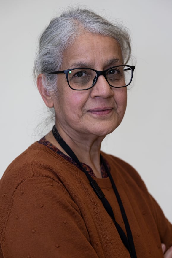 This headshot portrait photograph is of Farida, a brown woman in her 60s whose grey-white hair is tied back loosely so that part of it falls over her brow. She wears large dark-rimmed glasses and smiles in a composed way from the left of the image, facing the camera. She wears a tobacco brown knitted top with a round neck, over which hangs a dark staff lanyard. 