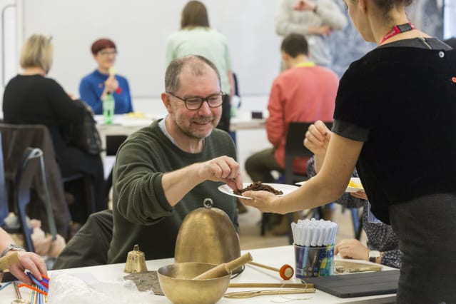 man leans forward whilst involved in aworkshop; the man is surrounded by other people, all of whom are using art materials in an atmosphere of enjoyable activity