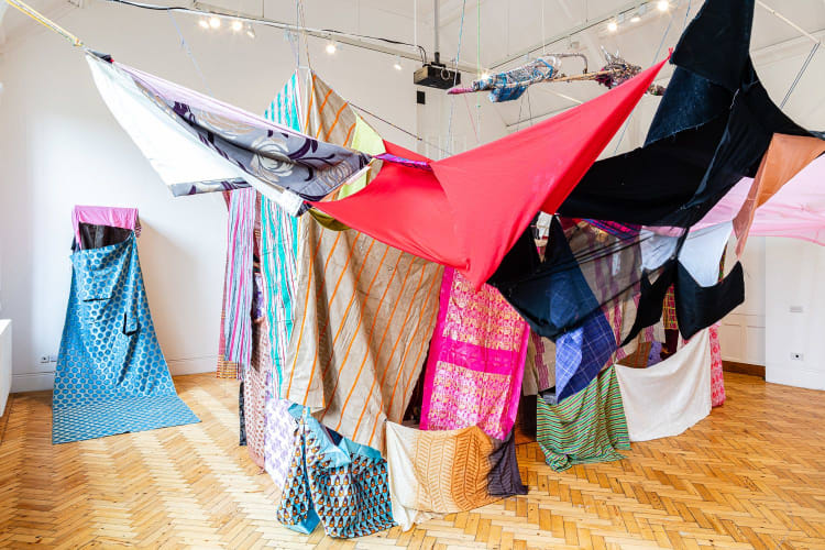 In the centre of a white, gallery space, individual rectangles of fabric are loosely strung together. They form a large tent-like structure; walls made from hanging cloth of varying patterns and a roof of colourful sheets, horizontally stretched from the corners of the room. On the far gallery wall, a piece of shiny blue fabric is attached about 2 metres off the wooden parquet flooring. It protrudes from the wall, inviting a visitor to come in and look out of the two window flaps, which have been cut into the fabric. 