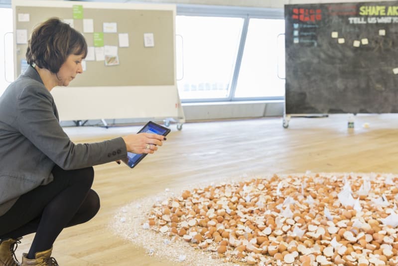 A woman crouches no the floor of a gallery space, holding an ipad and looking closely at an artwork - a wide, round, flat heap of eggshells on the floor