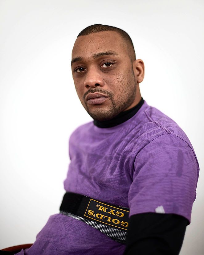 A black man is sat, slightly side on but with his head turned to face the camera. His expression is subtle, with a soft gaze. He wears a purple t-shirt and has short black hair.