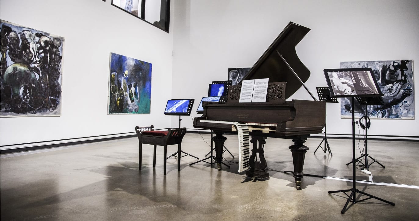 A black grand piano with half of the keys falling off sits in the middle of a very big, modern gallery space. There are screens on music stands around it and large, square abstract paintings on the walls behind it