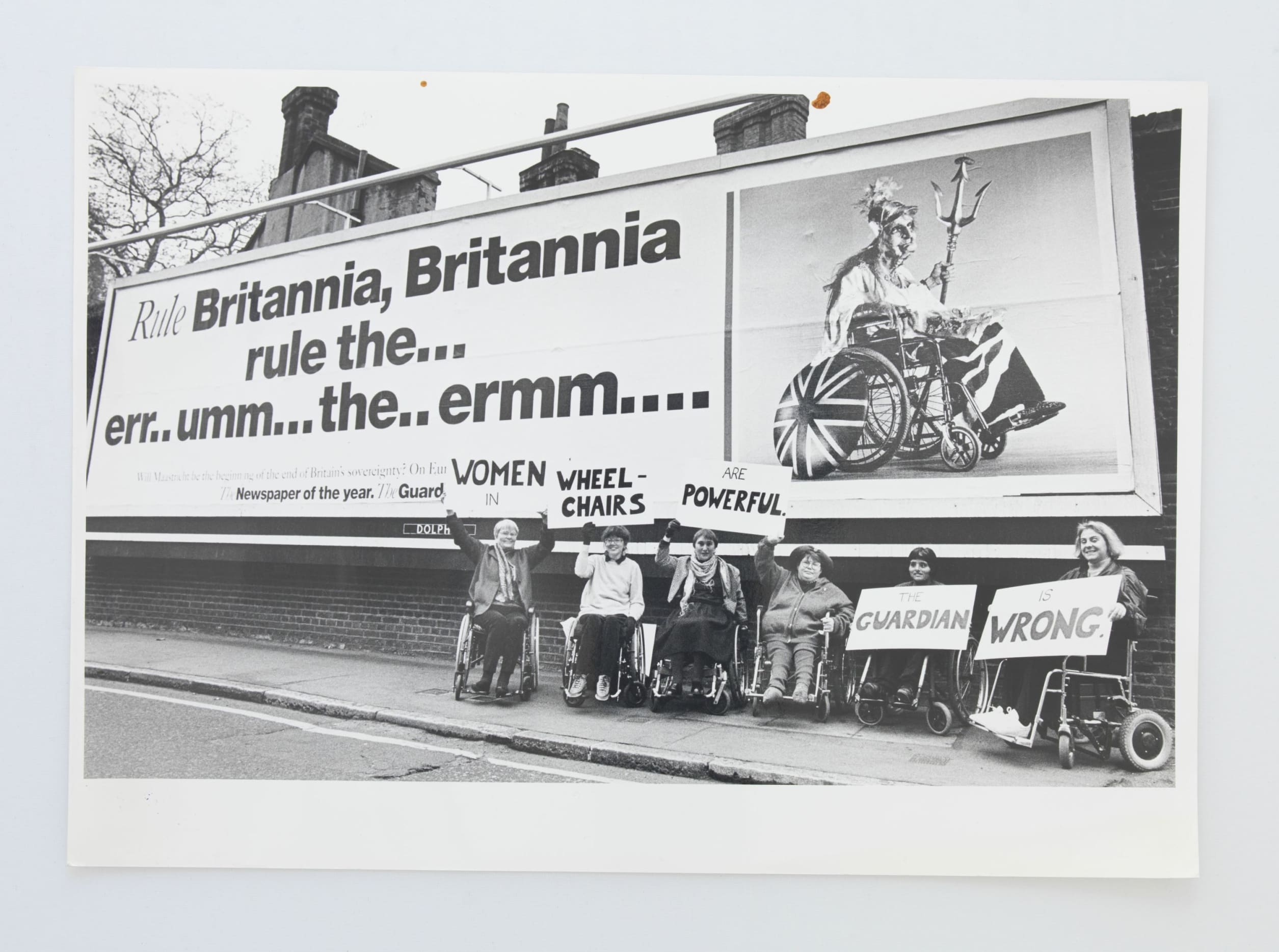 An old photograph of six female wheelchair users sitting underneath a billboard. The billboard is an advertisement for The Guardian newspaper when it won 