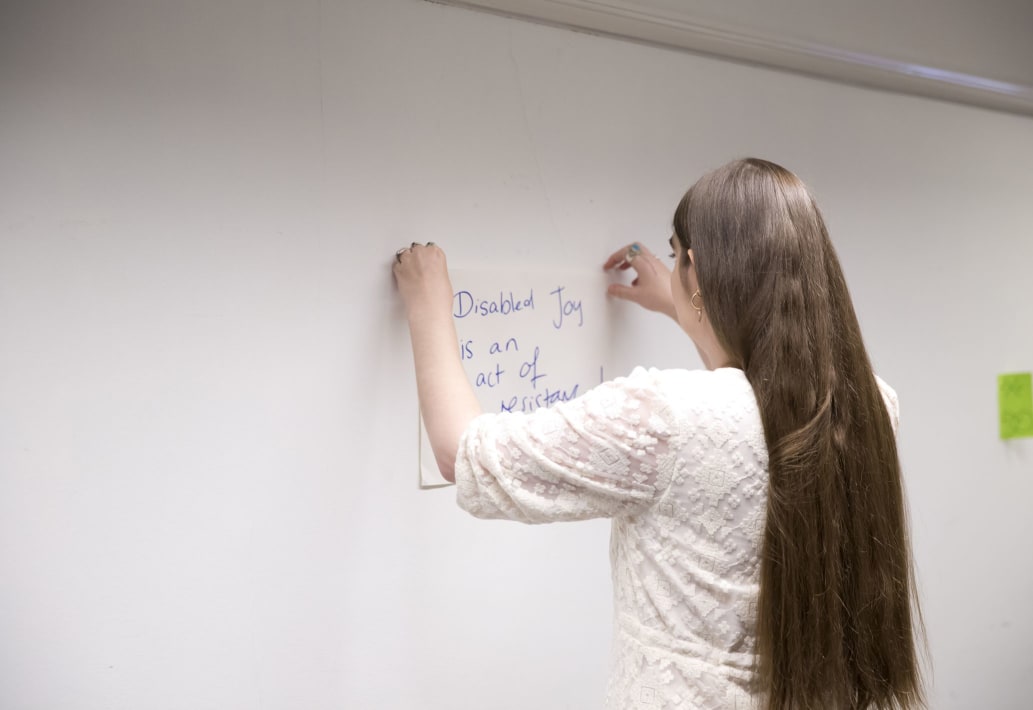 A photograph taken from behind of artist Charlie Fitz sticking a poster to the wall. Charlie is a young woman with very long, straight brunette hair. She is wearing a cream coloured lacy dress. In the photo, she is tacking a poster with hand written text that reads 