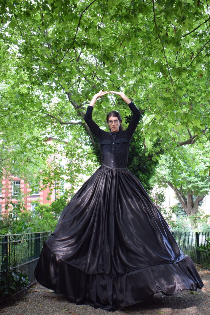 Abigail, a tall, white, trans woman with tied-back dark hair and glasses, is posing amongst a backdrop of trees and wild flowers, dressed in an extremely oversized black skirt suspended in place by a crinoline cage, whilst holding her arms in a ring 