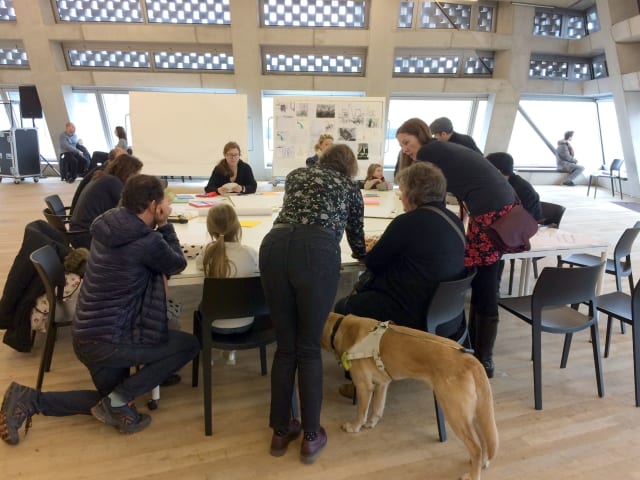 participants crowd around a table on which objects have been set out; an assistance dog indicates at least one of the participants is blind 