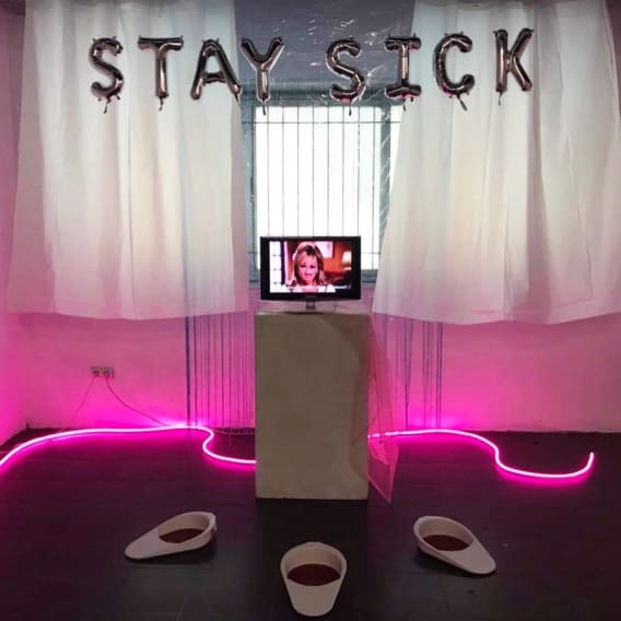 Photograph of part of an art installation. Along a plain white wall, two shower curtains are fixed, a long blue curtain to the right and a shorter transparent one centrally. To its left a much longer pink fabric is hung from higher up, partially drap