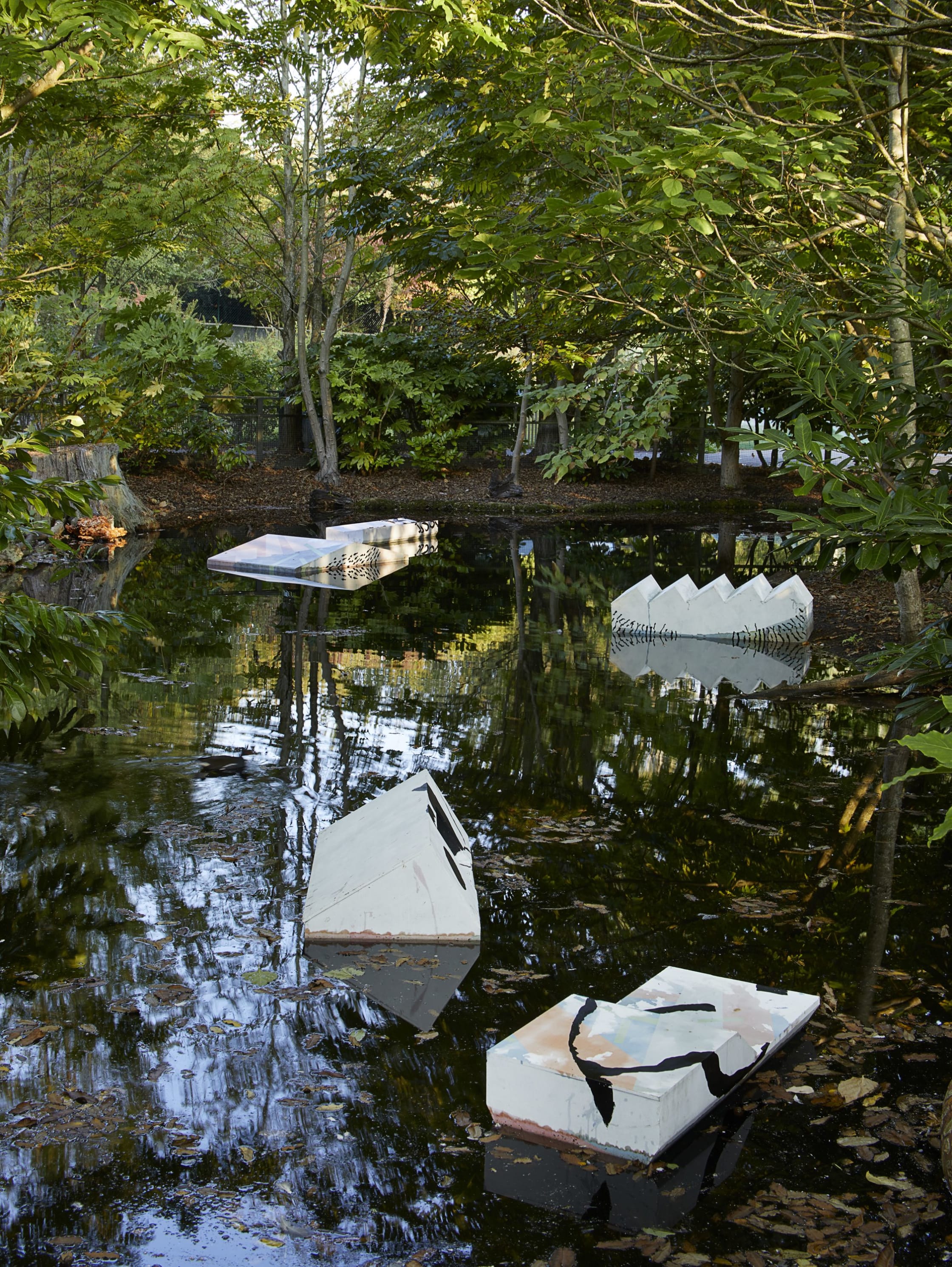 Photograph of an outdoor art installation. Green trees surround a small body of water, in which sit three sculptures. Each sculpture is white and resembles industrial aesthetics: stairs, for example.
