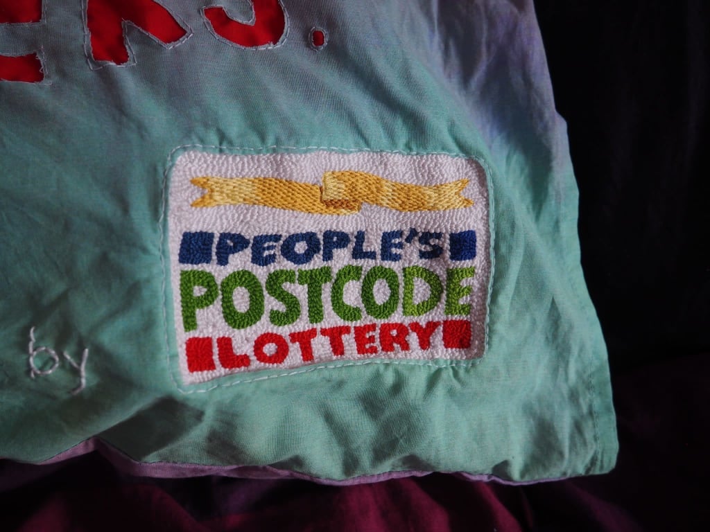 On a dark purple sheet of fabric is the corner of a pillow dyed with a fade of green to pink. A very finely detailed embroidered logo of ‘People’s Postcode Lottery’ covers the fabric with a fine white stitch following the edge of the logo.