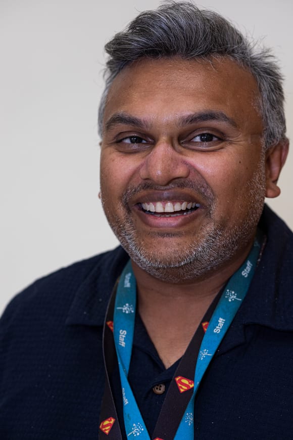 This portrait headshot of Sunnil shows a brown man in his 40s facing the camera from the right of the image with a broad smile that matches his jovial expression. He has greying dark hair with a fringe swept aside and a faint beard of greyish stubble. Hanging down before his dark shirt, whose top button is undone, are two staff lanyards, one pale blue and the other with superman logos across it.