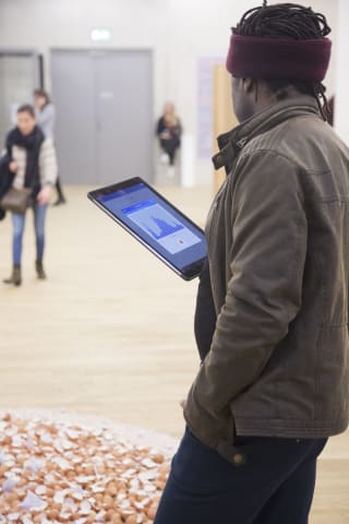 a man regards the ipad he is holding whilst standing before a floor installation of eggshells