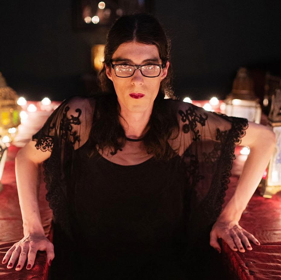 Abigail, a tall, white, trans woman with dark hair and glasses, is posing amongst a backdrop of candles warm light, dressed in black lace, holding herself up with both arms and faces the camera.