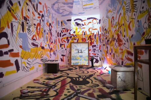 A photograph of a small white walled gallery space covered in blocks of coloured paint across the walls and floor. A mirror is stood at the far end of the room.