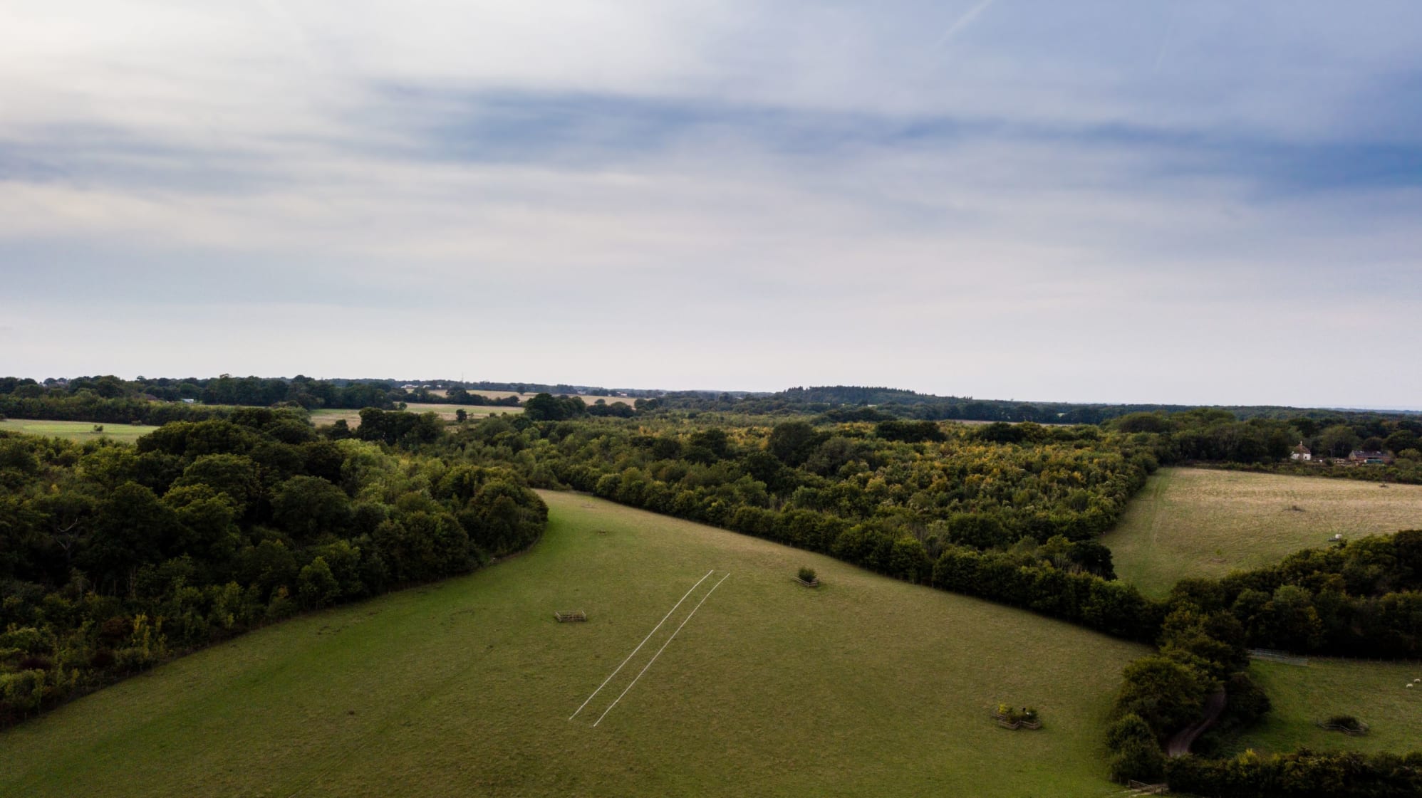 Aerial photograph of Annes artwork in its landscape. An expanse of green fields stretch out away from the camera. The sky blue and cloudy. On the cleared field below, are two white chalk lines placed in the grass.