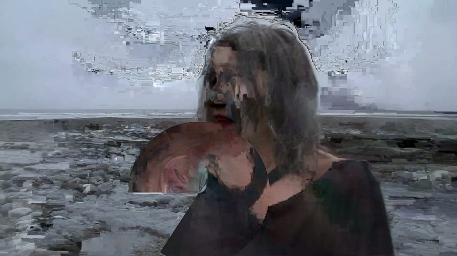 A still from a video. A woman with long grey hair stands close to the camera infront of a grey beach. Her face is pixelated and distorted, blurring her facial features.