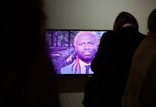 Photo of a large tv screen displaying a close of up Samiir Saunders. People are gathered around the screen.
