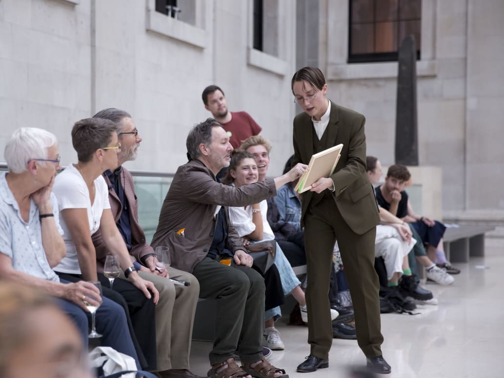 Photograph of an audience participation moment during a performance taking place in the Great Court of the British Museum. Many audience members are sat along a long silver bench. Behind them, the white stone walls of the court provide a backdrop to the performance. The performer, Dre