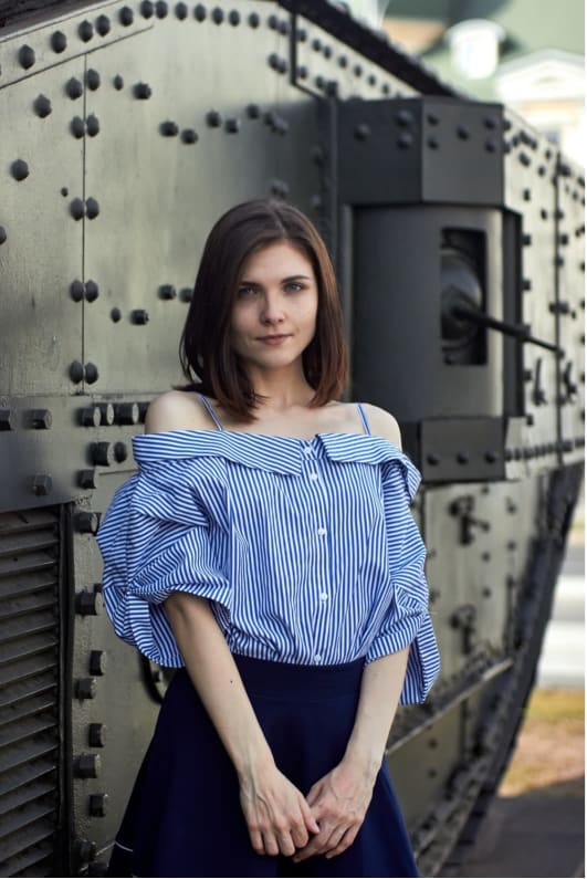 Anna Moshak, stands central to the image with long brown hair, wearing a striped blue shirt and blue skirt. She stands in front of a tank.