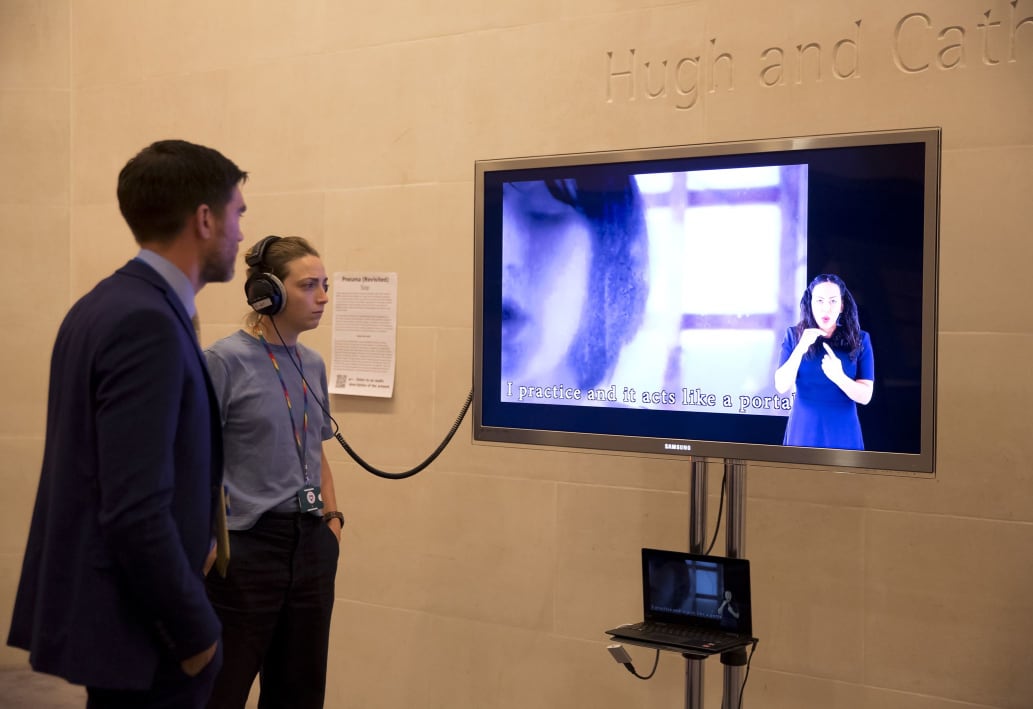 A photograph of two audience members, a man with a beard and a woman with short hair wearing a blue cropped jumper, stood in front of a TV screen. The woman is wearing headphones that connect to the screen. On the screen, an artist film by Sop plays. Sop