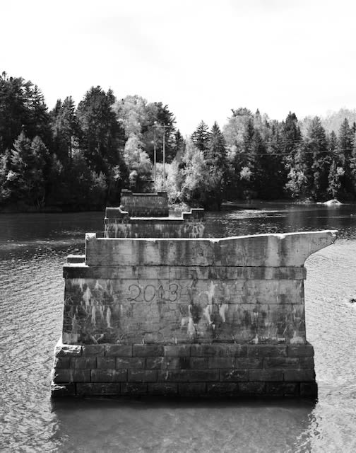 The derelict bridge in Canada that inspired Faes work. Image is black and white and shows a broken bridge across a small river behind which are trees.