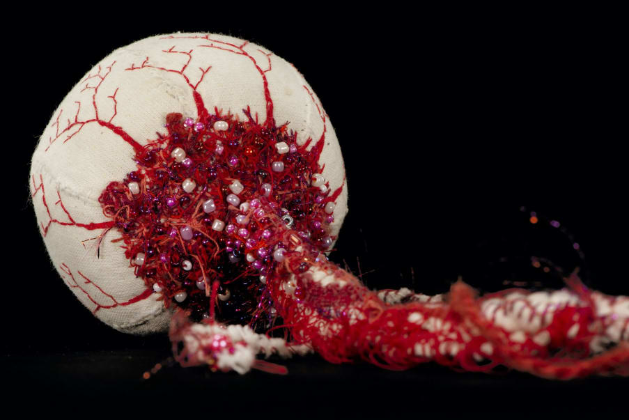 A close up photograph of a delicately and colourfully embroidered model of a human eye. The iris and pupil are facing away from the camera, so the audience is shown the intricate vessels and veins, crafted from rich red thread and adorned with matching small beads.