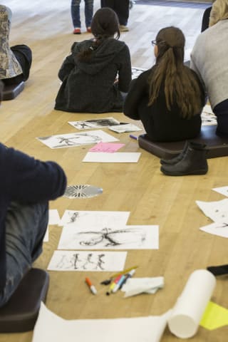 Young people sitting with their backs to us, and looking up intently in a worskshop setting. Sketches on paper are on the floor, suggesting they are learning how to draw with charcoal or pencil of the same age, both drawing thoughtfully whilst kneeling
