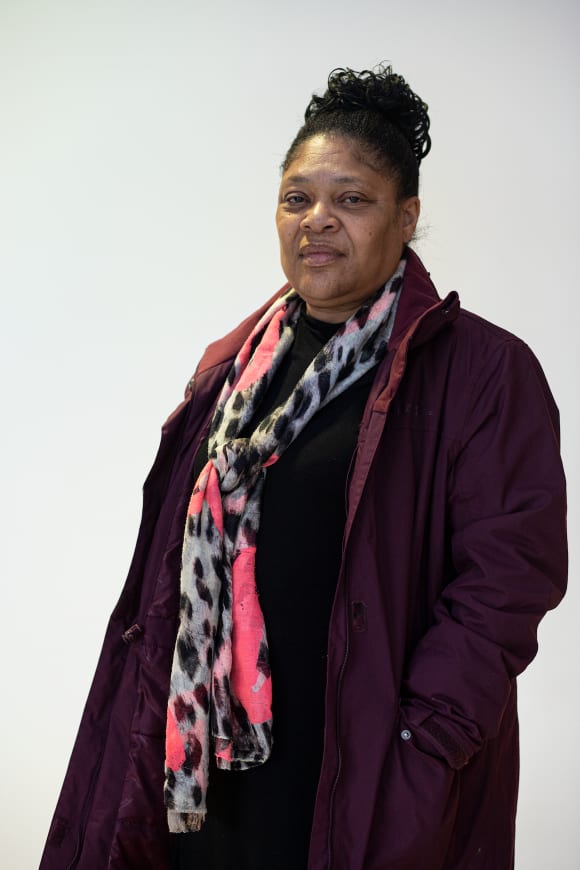 This portrait photograph is of Maxine. This is almost a full length portrait of a black woman in her 60s, with finely braided dark hair piled up in a neat bun, and who wears a dark purple winter coat and black and white spot-printed scarf with vibrant markings of pink. She has a calm, even expression and faces the camera from the right side of the image, her head turned slightly to the left. Her arms are at her sides; on the left side we find her hand buried deep in her pocket. 