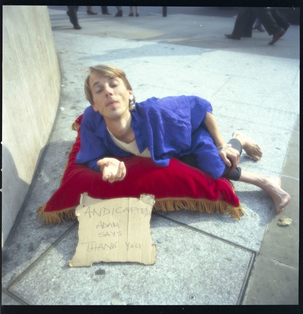 A slim white man in a blue robe is laying on a red velvet cushion on a pavement with his hand outstretched and eyes closed. A cardboard sign in front of him reads Andicapped Adam says thank you