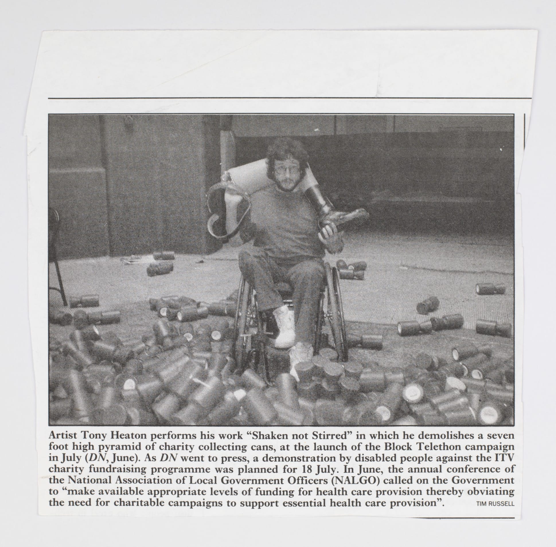Black and white newspaper image of a young white man sitting in a wheelchair with a prosthetic leg slung over his shoulders. He is surrounded by knocked-over charity collecting cans