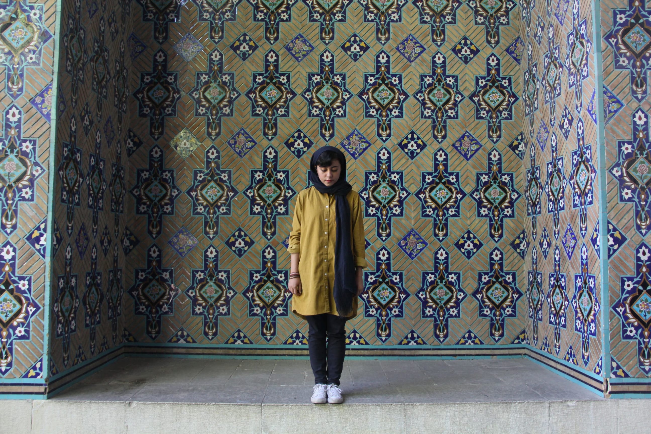 A woman stands in a large tiled alcove. She wears white trainers, black trousers, mustard button down shirtdress and navy head scarf with one end folded over her shoulder and the other trailing down her chest. Her head is tilted forwards as she looks