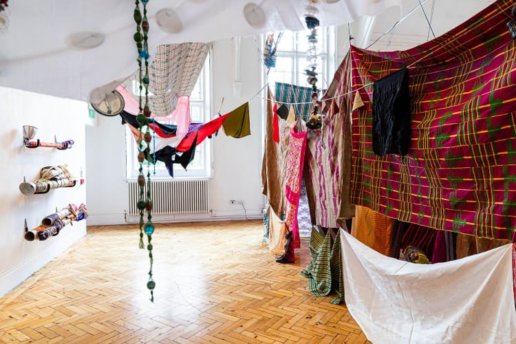 A view of a gallery installation framed along the top edge by a piece of white cotton fabric which we appear to be peering from underneath. Two strings of glass beading hang in the foreground from somewhere above, just out of shot.  Behind this, stretching from the right corner and into the middle of the room is a wall of individual rectangles of colourful fabric strung loosely together. On the left is a white gallery wall on which three sculptural forms are attached, evenly spaced and stacked horizontally. They are abstract, long and tubular in form. Wrapped partially in colourful wool. On the far wall, light spills into the room and onto the wooden parquet flooring from two tall windows. 