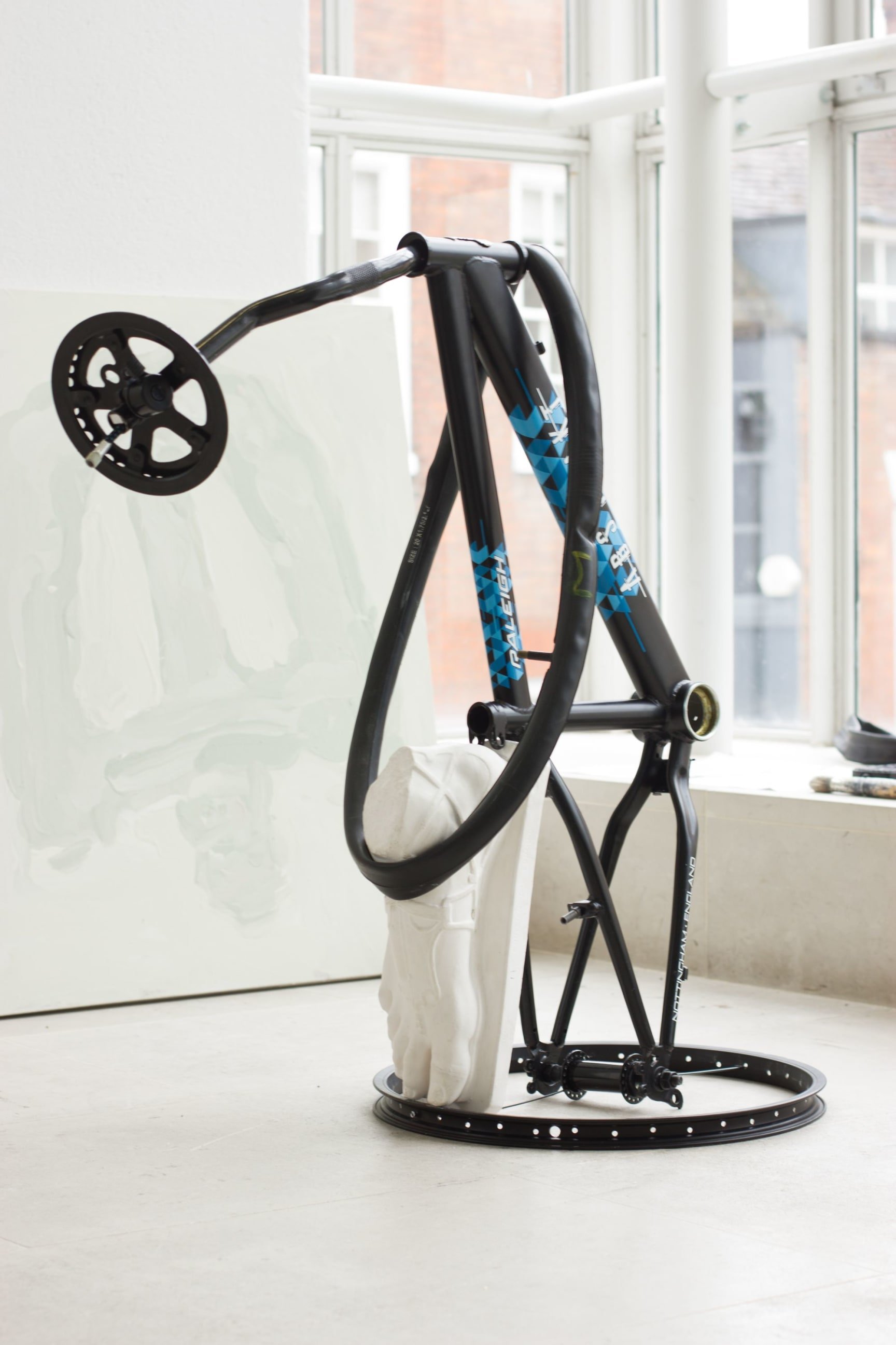 Abstract sculpture on the floor of a studio space in front of bright windows. It appears to be made of bicycle parts and a plaster foot in the style of a classical sculpture.