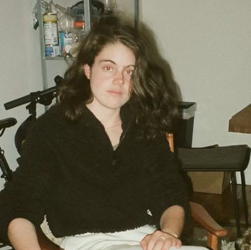 A photograph of Sophie, with mid-length brown wavy hair and wearing a black button-up shirt, sitting in a chair.