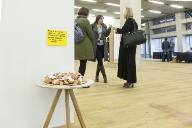 in the foreground, under a curatorial notice, a plate of eggshells acts as a model for touch exploring the larger eggshell installation it sits adjacent to, although the installation is out of the picture; in the background three woman are talking