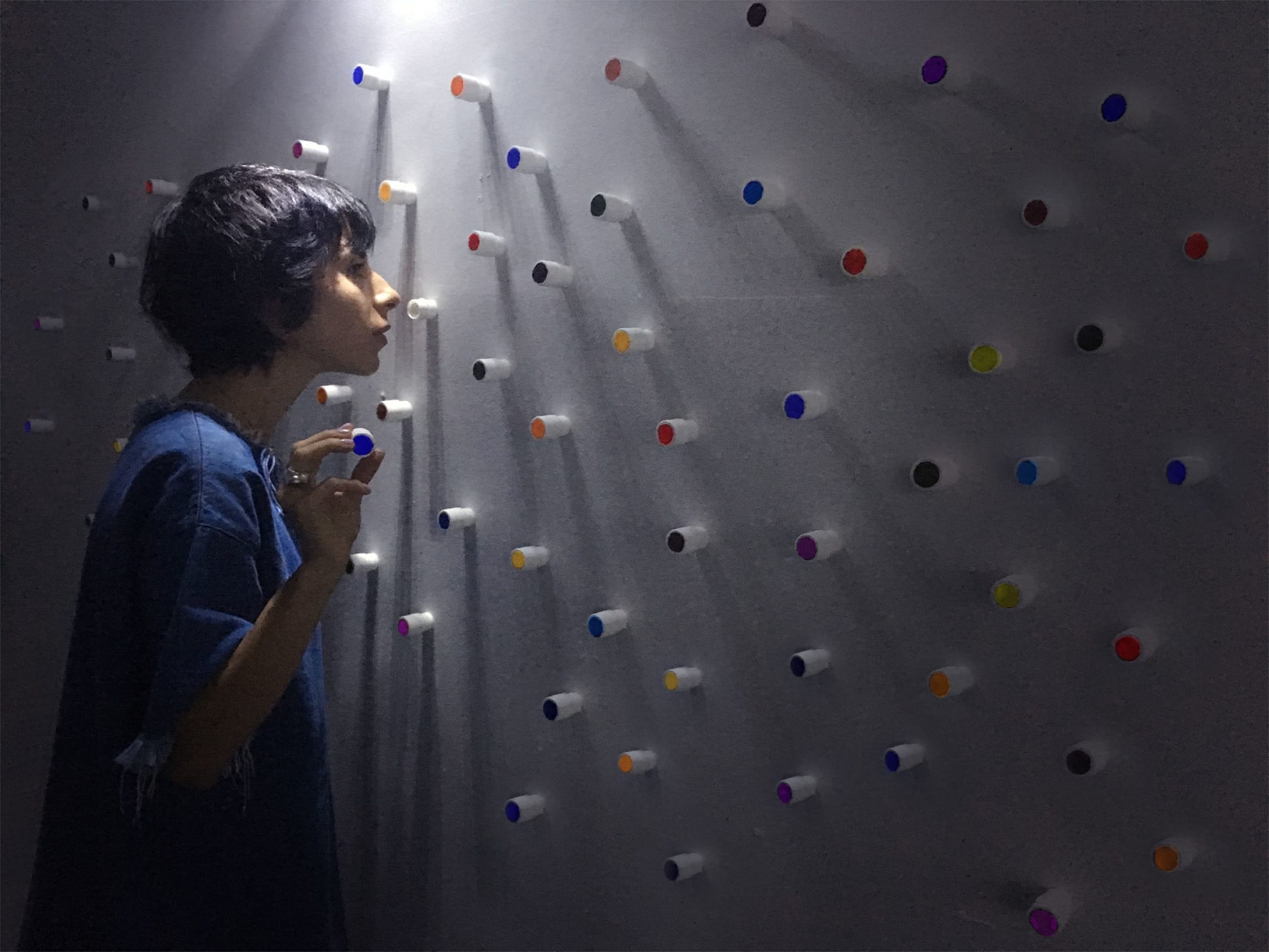A woman with olive skin and short black hair stands in a dark room with her face and hands close to a perspex wall containing many multicoloured tubes at regular intervals. She is smelling the tubes.